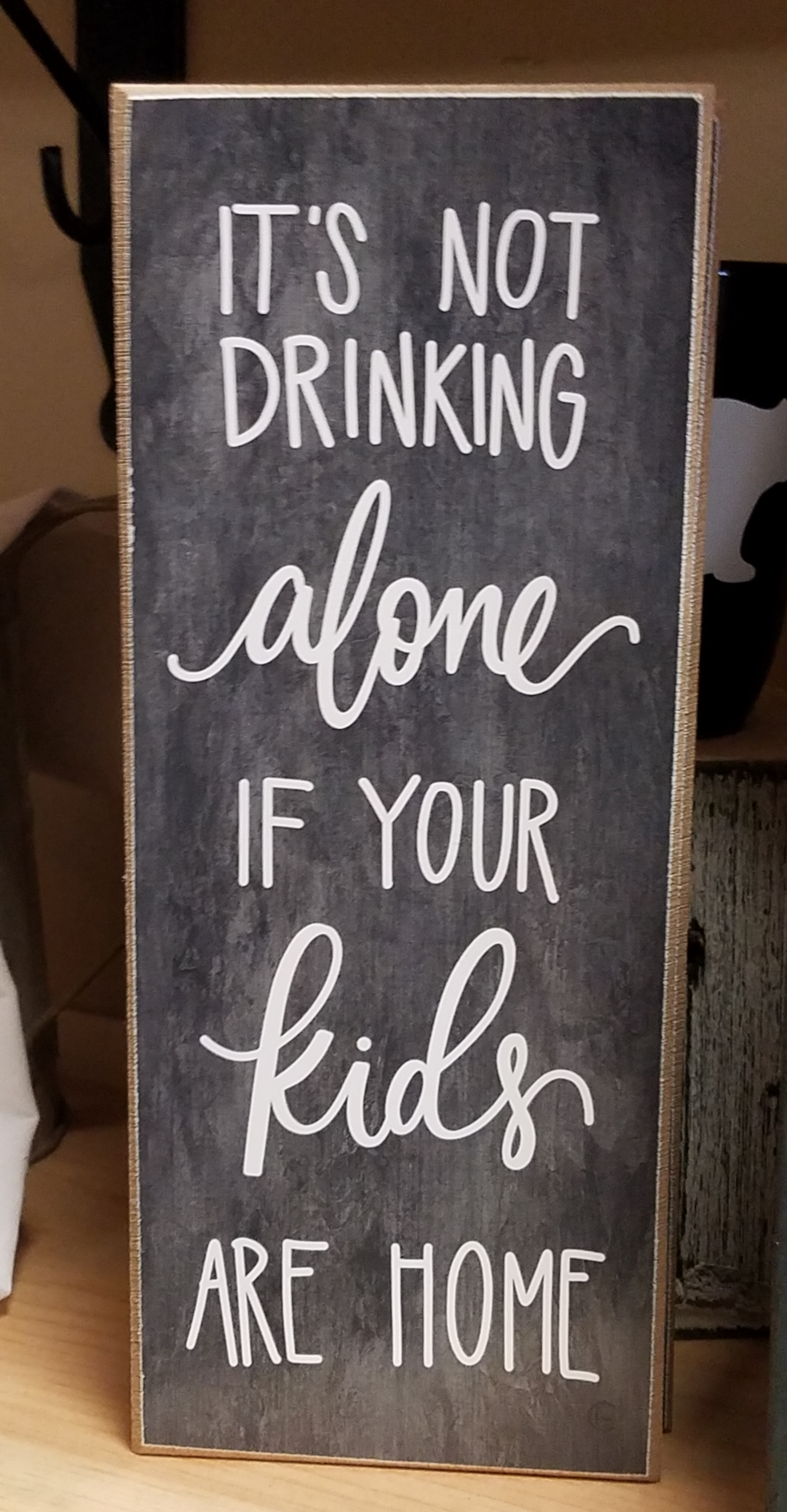 Dark grey background (chalkboard style) that reads "It's not drinking alone if your kids are home" in white. The 10X4X1 size makes it perfect to display on table-top, shelf or mantle.