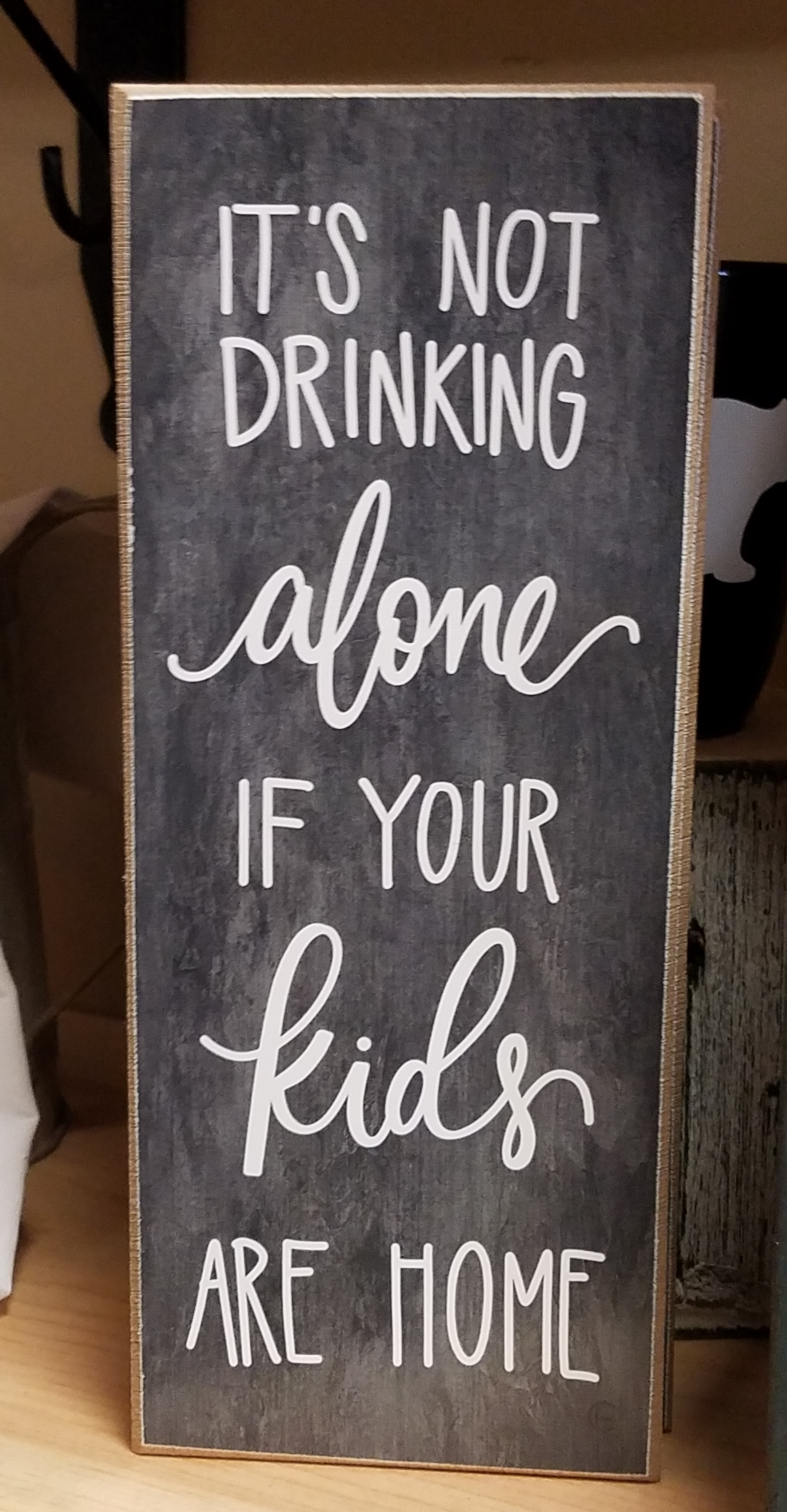 Dark grey background (chalkboard style) that reads "It's not drinking alone if your kids are home" in white.  The 10X4X1 size makes it perfect to display on table-top, shelf or mantle.